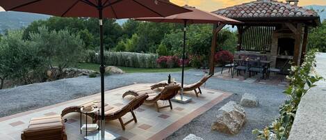 An outdoor patio with lounge chairs and a pergola, surrounded by a gravel path and greenery.