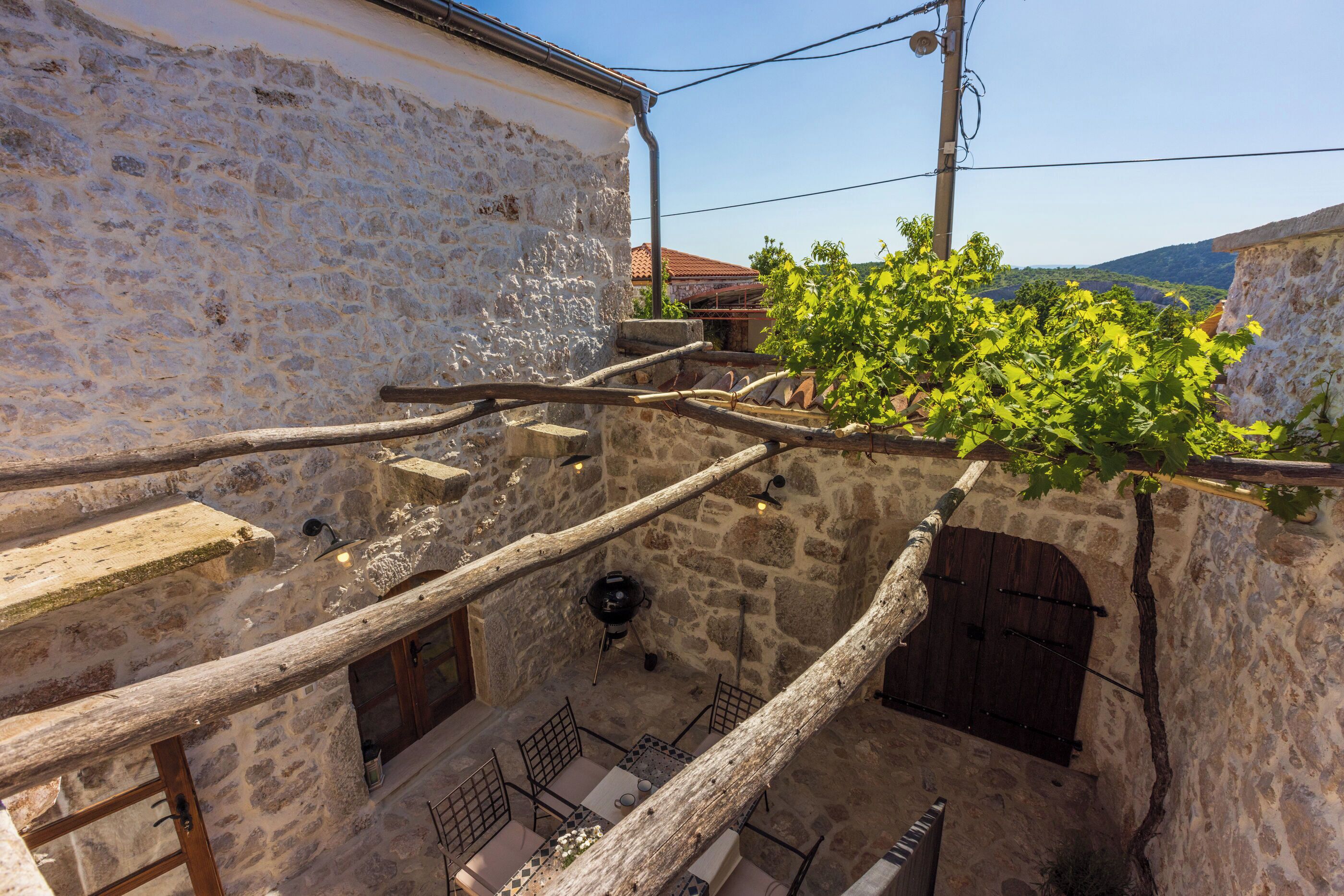 Une cour pavée de pierres, agrémentée d’une pergola en bois, d’une petite table avec des chaises et offrant une vue sur une colline.