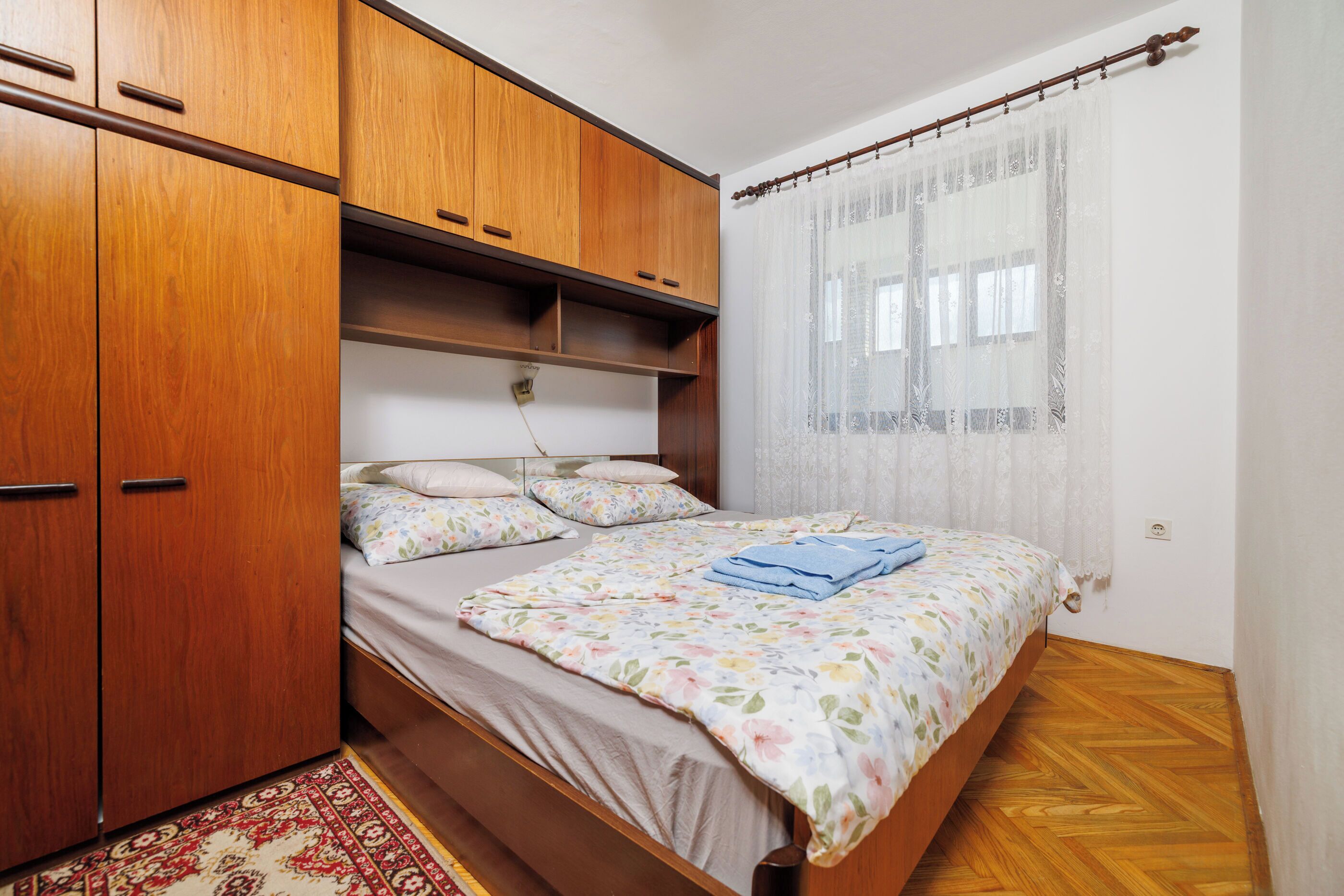 A bedroom with a bed, wooden wardrobe, and a window with sheer curtains.