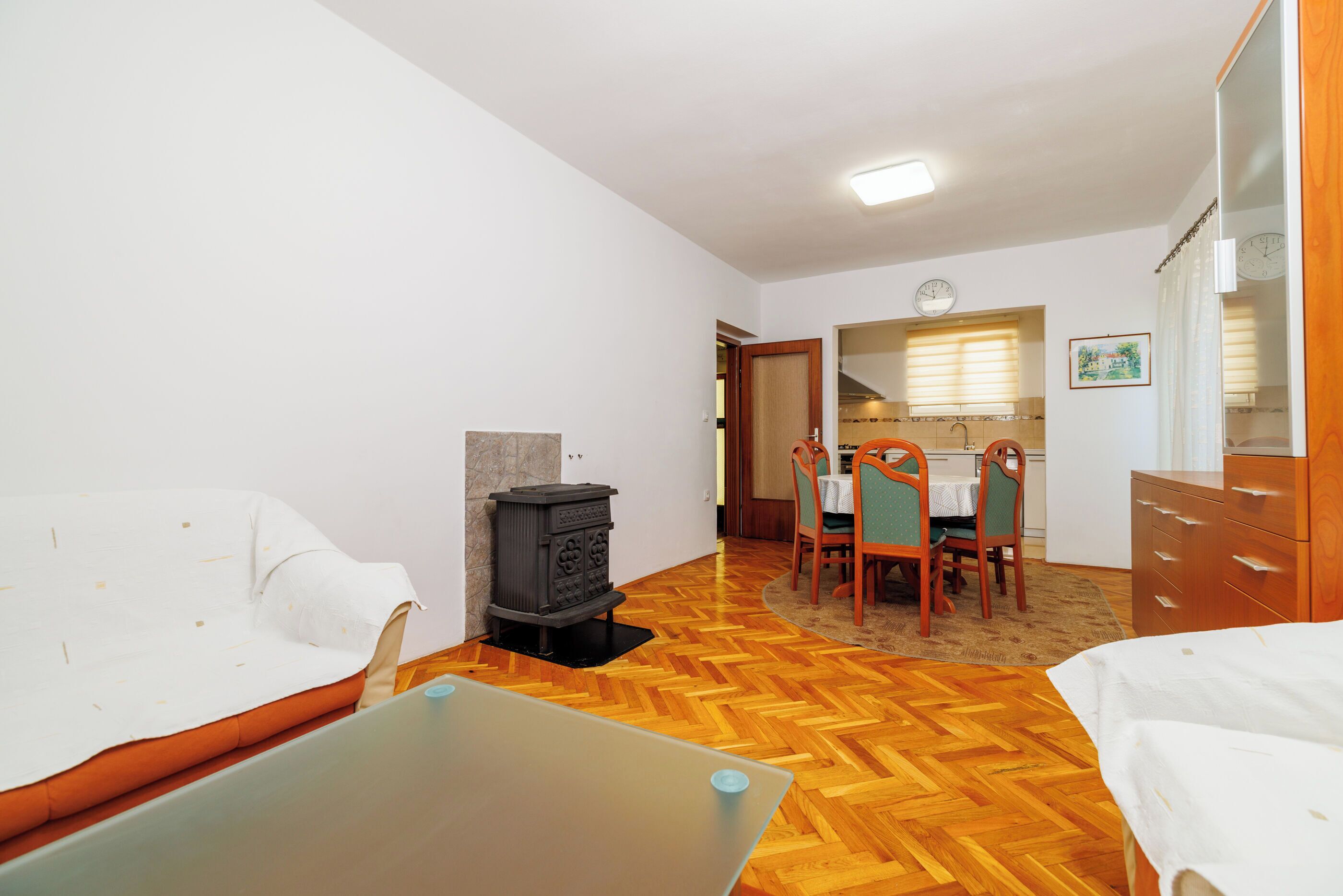 Une pièce comprenant un canapé, une petite table, un parquet en bois disposé en point de Hongrie, un poêle, un coin repas avec des chaises et une table, ainsi qu’une commode en bois.
