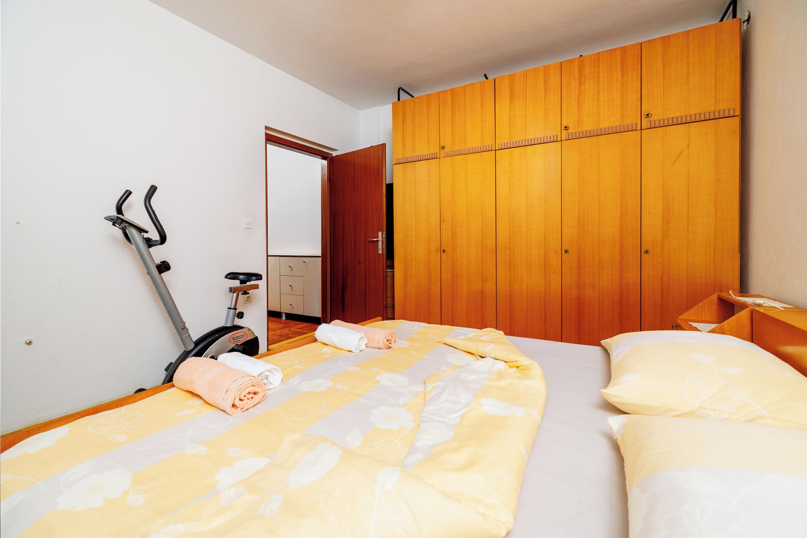 A bedroom with a bed, a wooden wardrobe, and a stationary exercise bike.