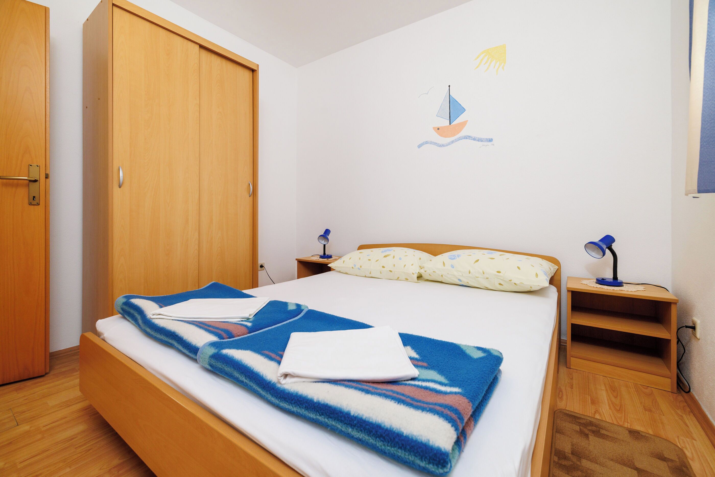 A neatly made bed with a blue and white blanket, a wooden wardrobe, a nightstand with a lamp, and a wall decoration of a sailboat.