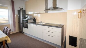 A modern kitchen with a stainless steel refrigerator, microwave, oven, and a gas stove. There is a window with a curtain, and a dining area with a table and chairs. - Apartments Vesna (Novi Vinodolski)