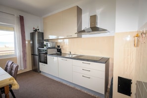 A modern kitchen with a stainless steel refrigerator, microwave, oven, and a gas stove. There is a window with a curtain, and a dining area with a table and chairs. - Apartments Vesna (Novi Vinodolski)