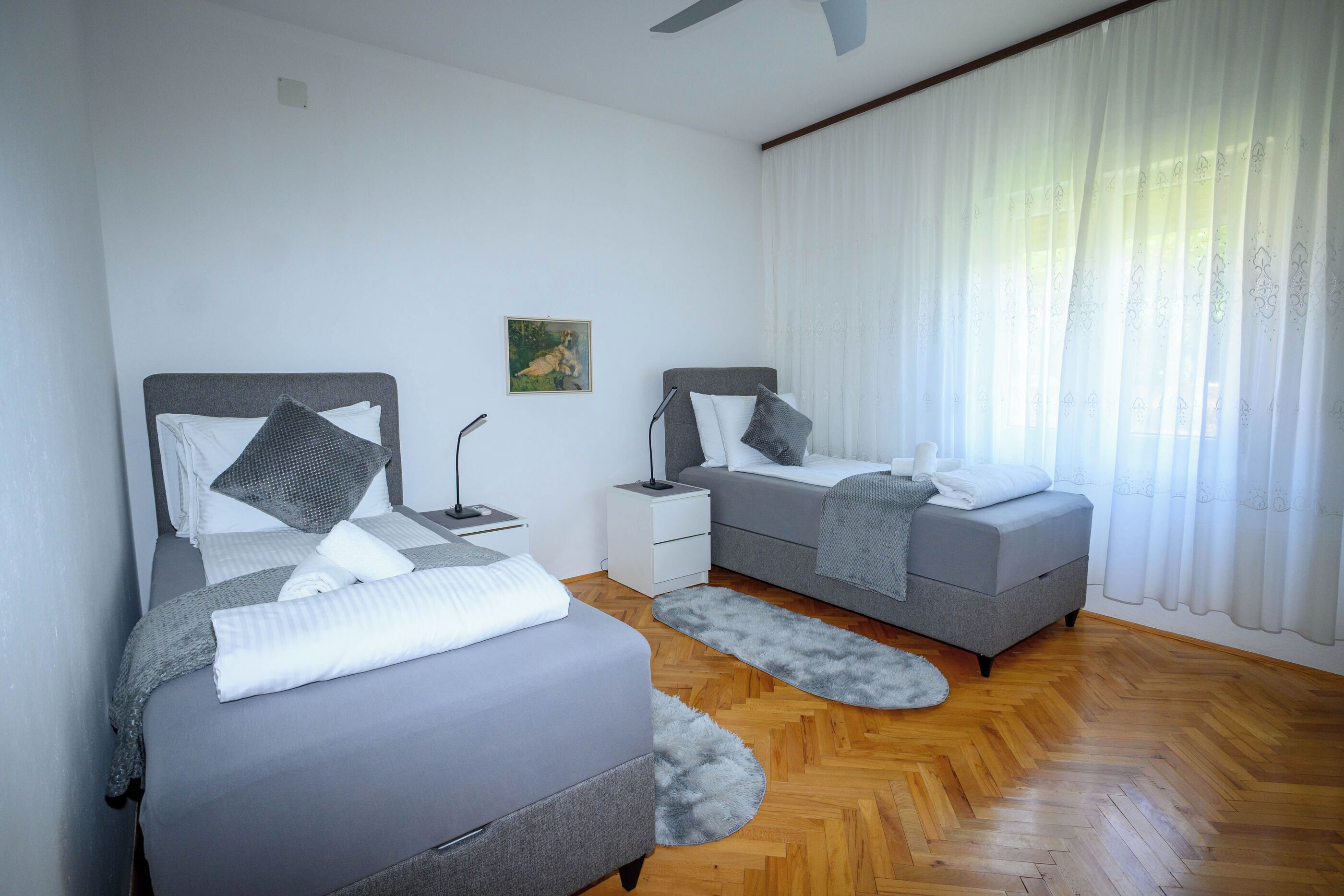 A modern bedroom with two beds, a nightstand, a lamp, and a painting on the wall.