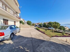 A residential street with a white car parked on the left, a stone pathway, and houses with balconies.