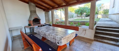 An outdoor patio with a wooden table and benches, a brick oven, and a view of a garden.