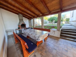 An outdoor patio with a wooden table and benches, a brick oven, and a view of a garden.