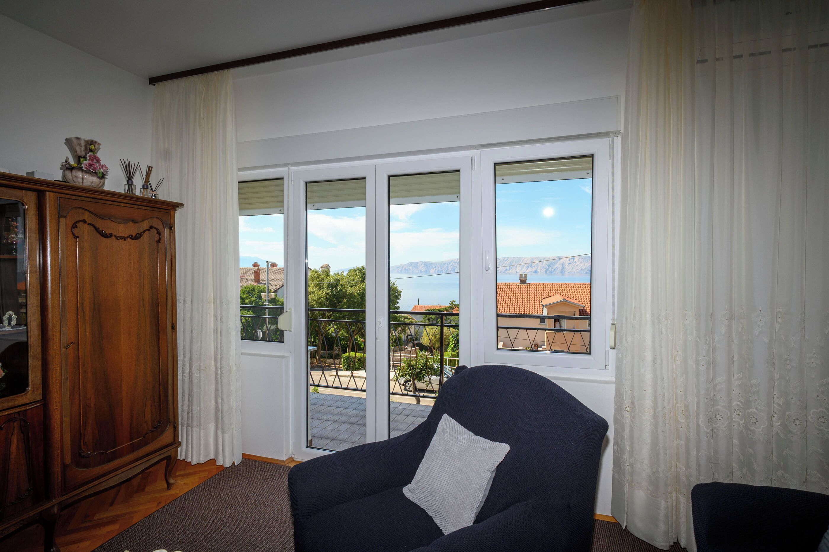 Una habitación con un sillón oscuro, un aparador de madera y vistas al exterior por una puerta corrediza de vidrio.