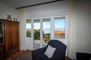 Une pièce avec un fauteuil sombre, un meuble en bois et une vue sur l’extérieur, accessible par une porte-fenêtre coulissante.