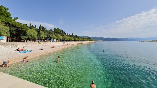 A beach with clear water, people swimming and sunbathing, and a row of trees and buildings along the shore. - Apartments Vesna (Novi Vinodolski)