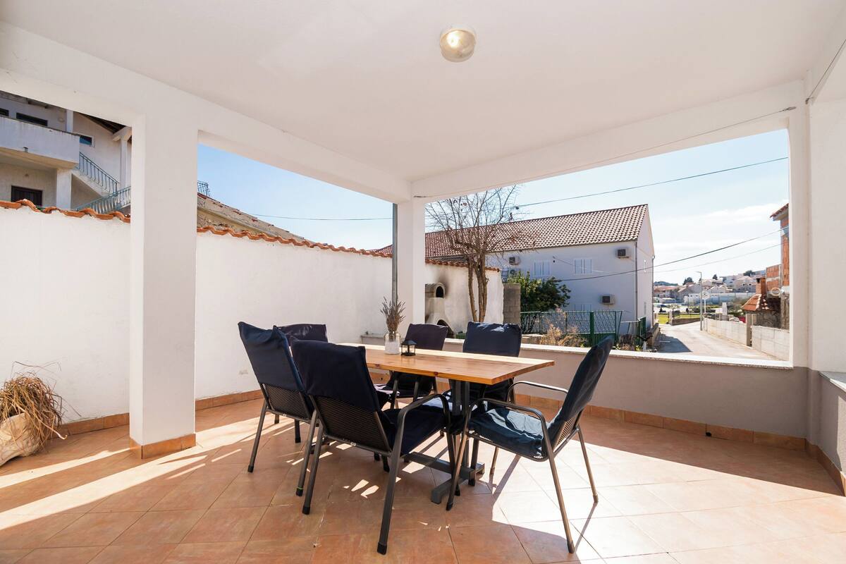 Une terrasse avec une table et des chaises en bois, une vue sur un bâtiment avec un toit en tuiles et un ciel dégagé.