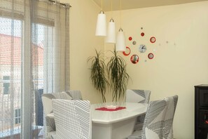 A dining area with a white table, patterned chairs, hanging pendant lights, a potted plant, and wall decorations including clocks. - Apartments Anita Tribunj (Tribunj)