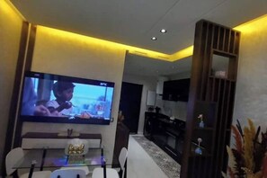 Un salon moderne avec un téléviseur à écran plat fixé au mur, un coin repas avec une table en verre et une cuisine dotée de meubles de couleur sombre.