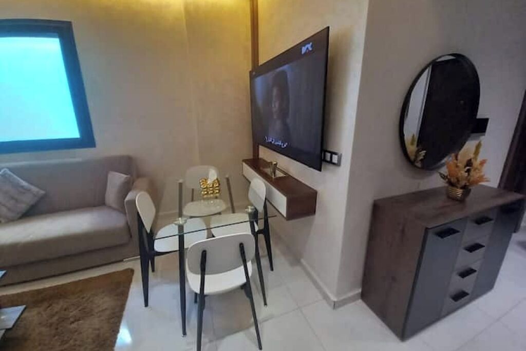 A modern living room with a sofa, a glass dining table, and chairs. A flat-screen TV is mounted on the wall, and there is a small cabinet with a mirror.