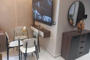 Un espace repas moderne avec un téléviseur fixé au mur, une table en verre, des chaises blanches et un meuble en bois doté de tiroirs.