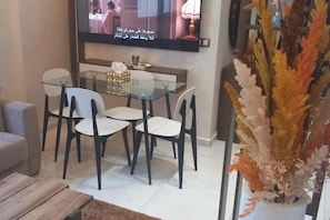 A dining area with a glass table and chairs, a flat-screen TV mounted on the wall, and a decorative plant in the foreground. - Welcome home! (Kénitra)