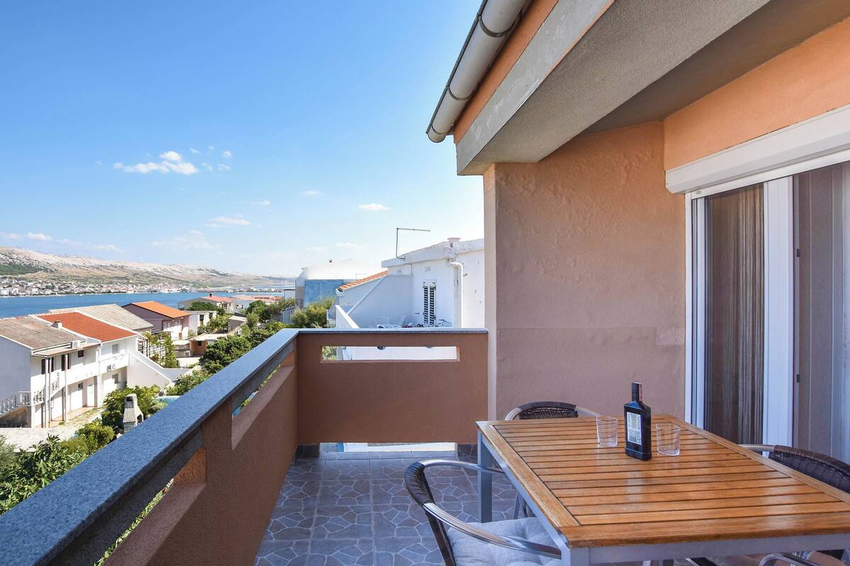 Un balcon avec une table et des chaises en bois, une bouteille, et une vue sur une ville côtière.