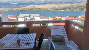 Ein Balkon mit Tisch und Liegestuhl, mit Blick auf einen See und eine Stadt mit roten Dächern.