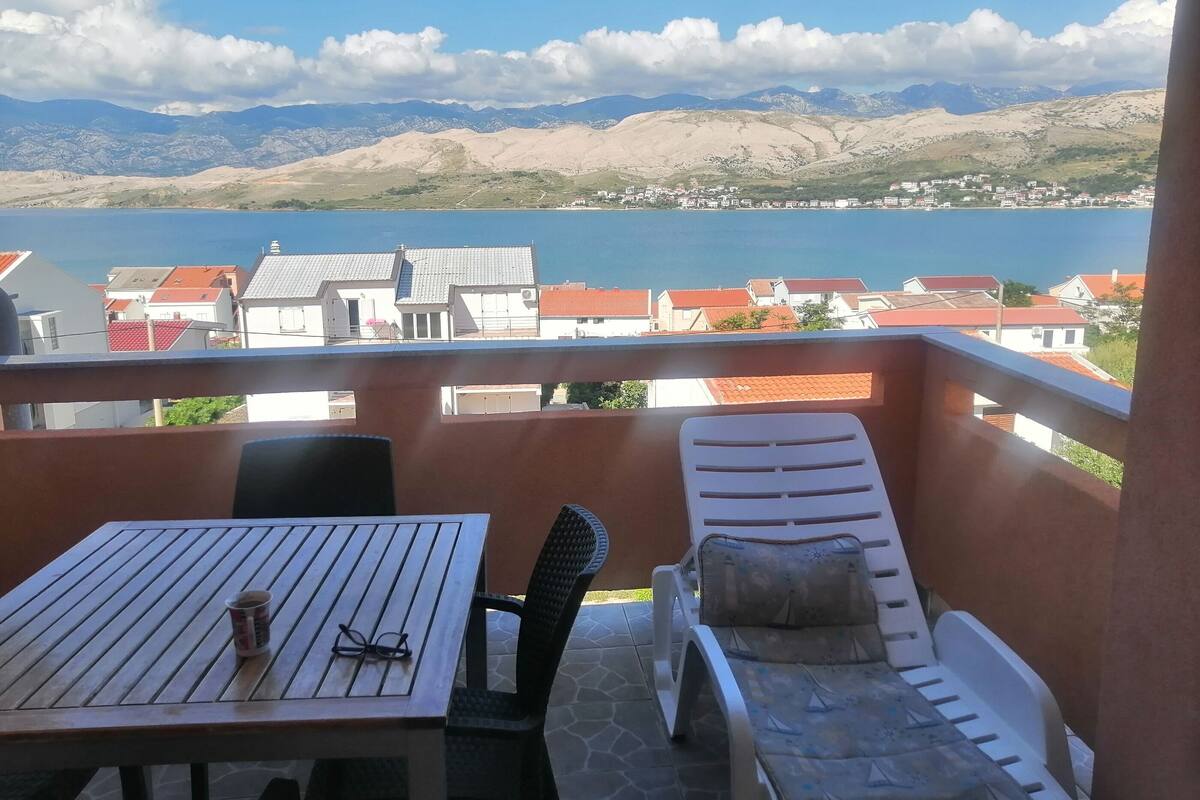 Un balcon avec une table et un fauteuil, donnant sur un lac et sur une ville aux bâtiments aux toits rouges.