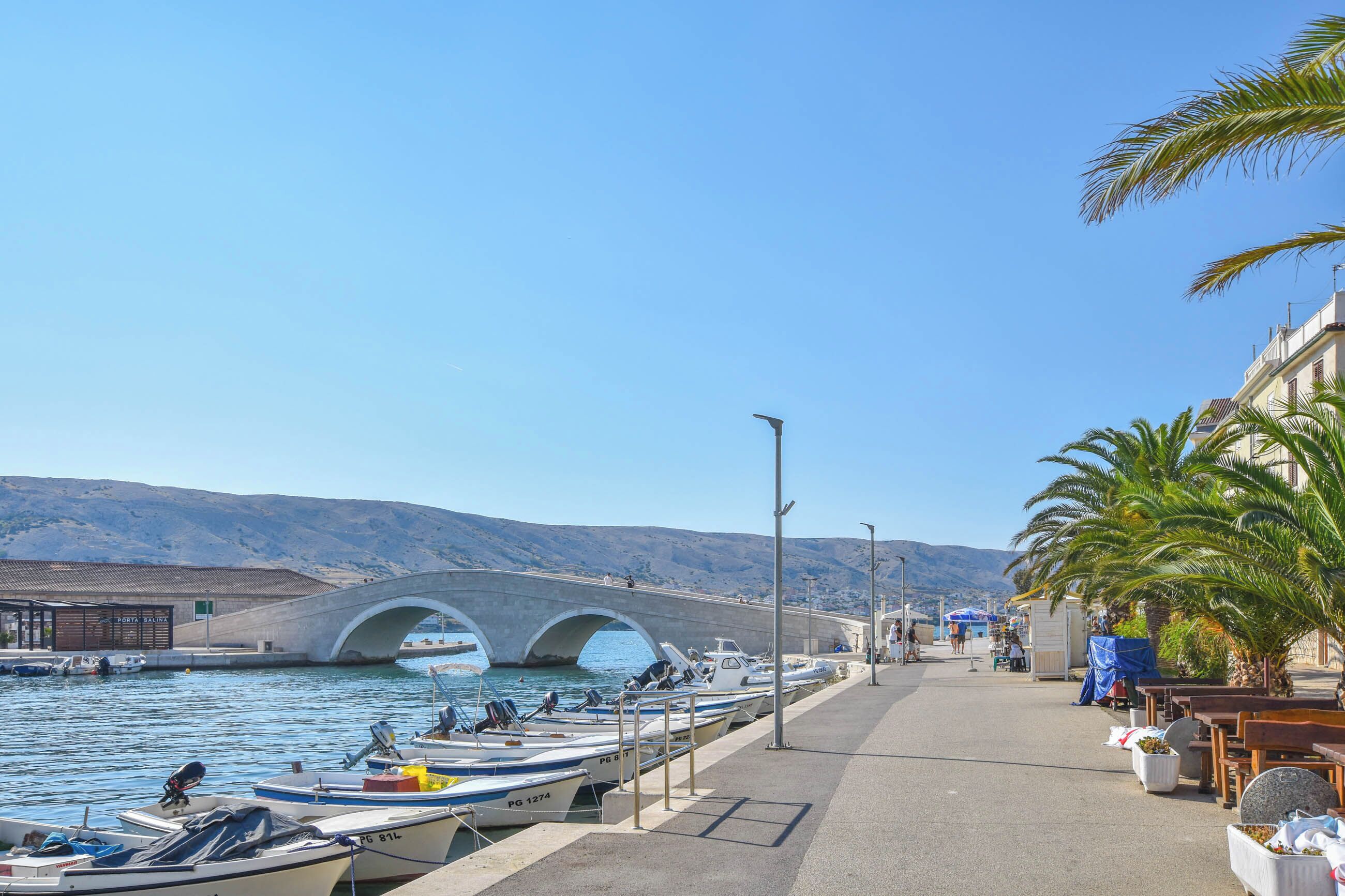 A coastal marina with boats docked, a pedestrian walkway, and a bridge in the background.