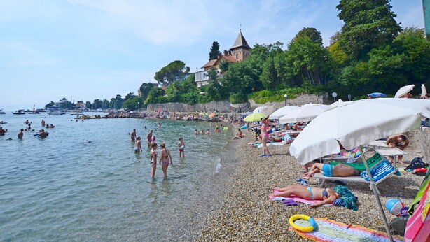 Ein Strand mit badenden und sonnenden Menschen, Sonnenschirmen und einem historischen Gebäude im Hintergrund.