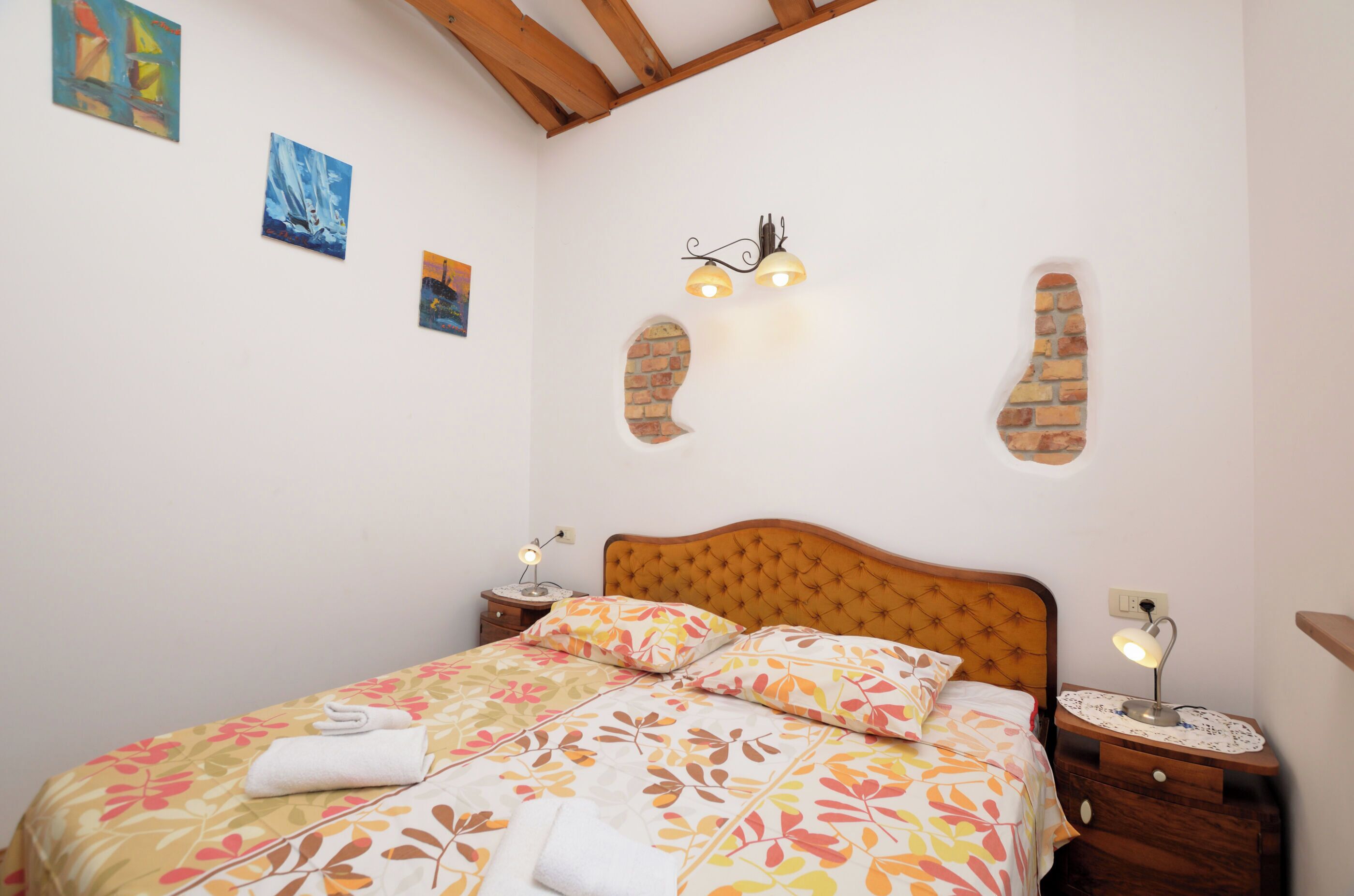 Une chambre à coucher comprenant un lit, deux tables de chevet, un luminaire fixé au mur et une petite fenêtre ornée d’un motif en briques.