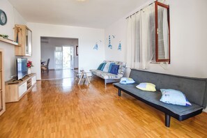A spacious living room with a wooden floor, a grey sofa, a TV, and a dining area in the background. - House Marko (Pasman)