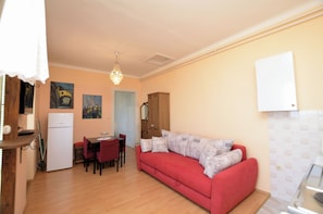 Ein gemütlicher Wohnbereich mit einem roten Sofa, einem Esstisch mit Stühlen, einer kleinen Küchenzeile mit einem Kühlschrank und einem Holzschrank.