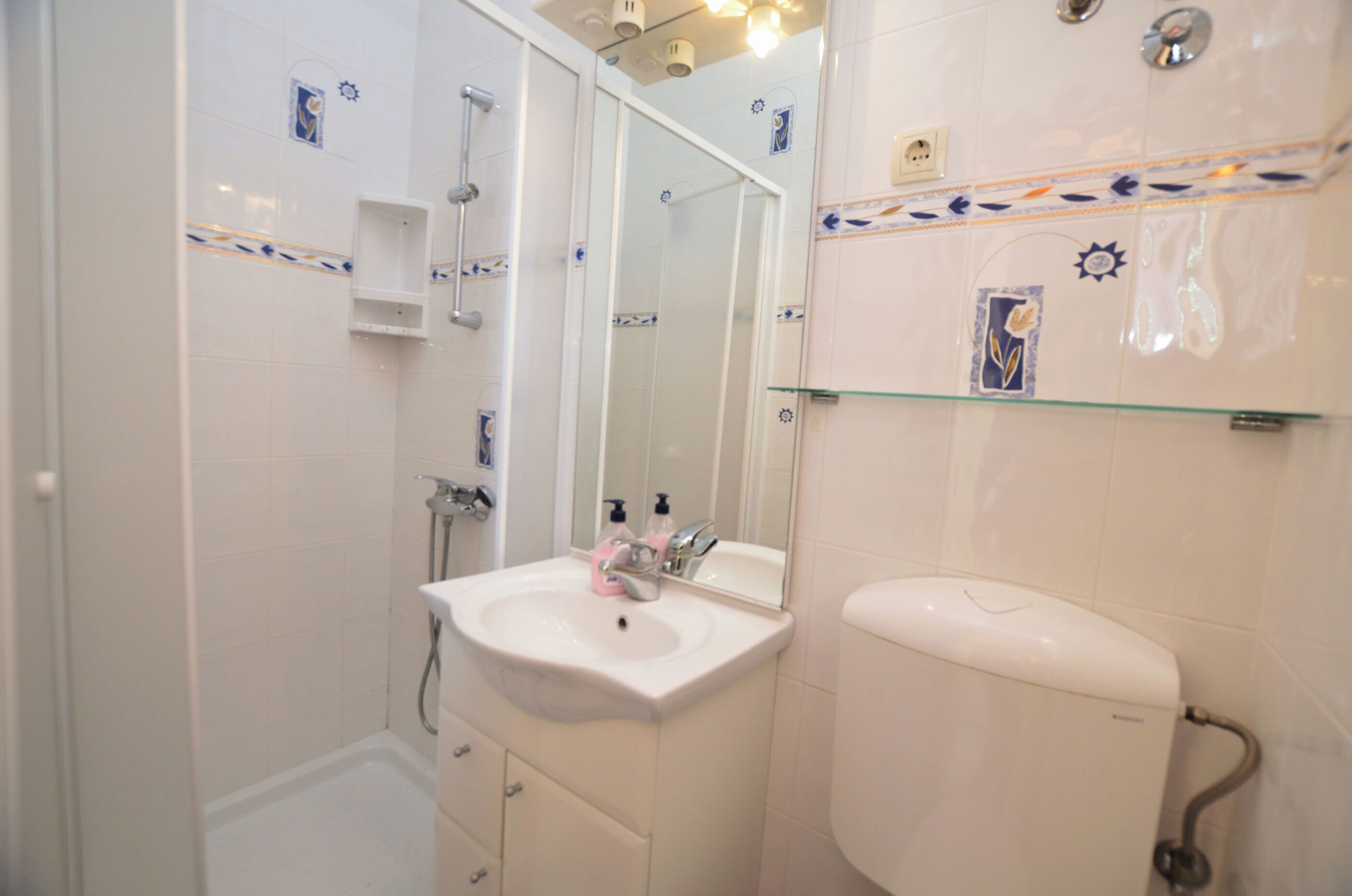 Une salle de bain avec un lavabo blanc, un miroir, une étagère en verre et des toilettes.