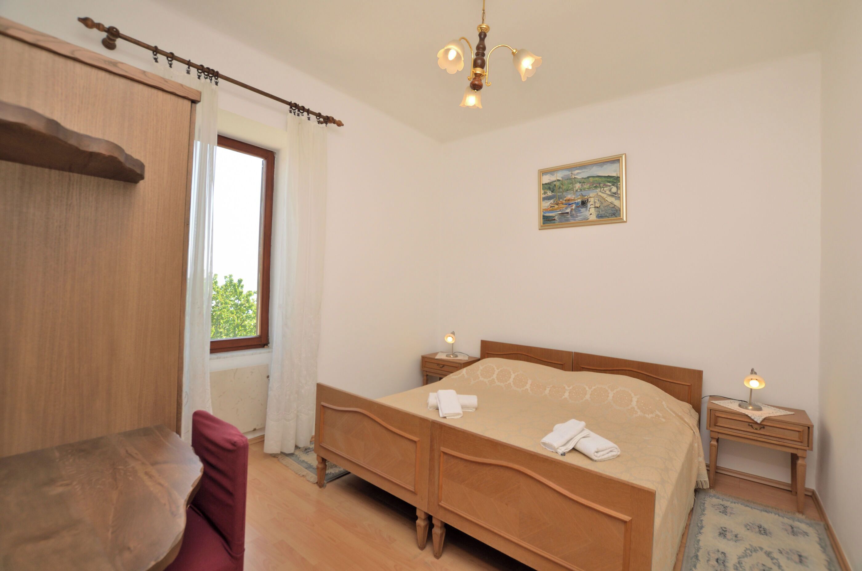 Ein Schlafzimmer mit einem hölzernen Bett, Nachttischen, einem Fenster mit Vorhängen, einem Gemälde an der Wand und einer Deckenleuchte.