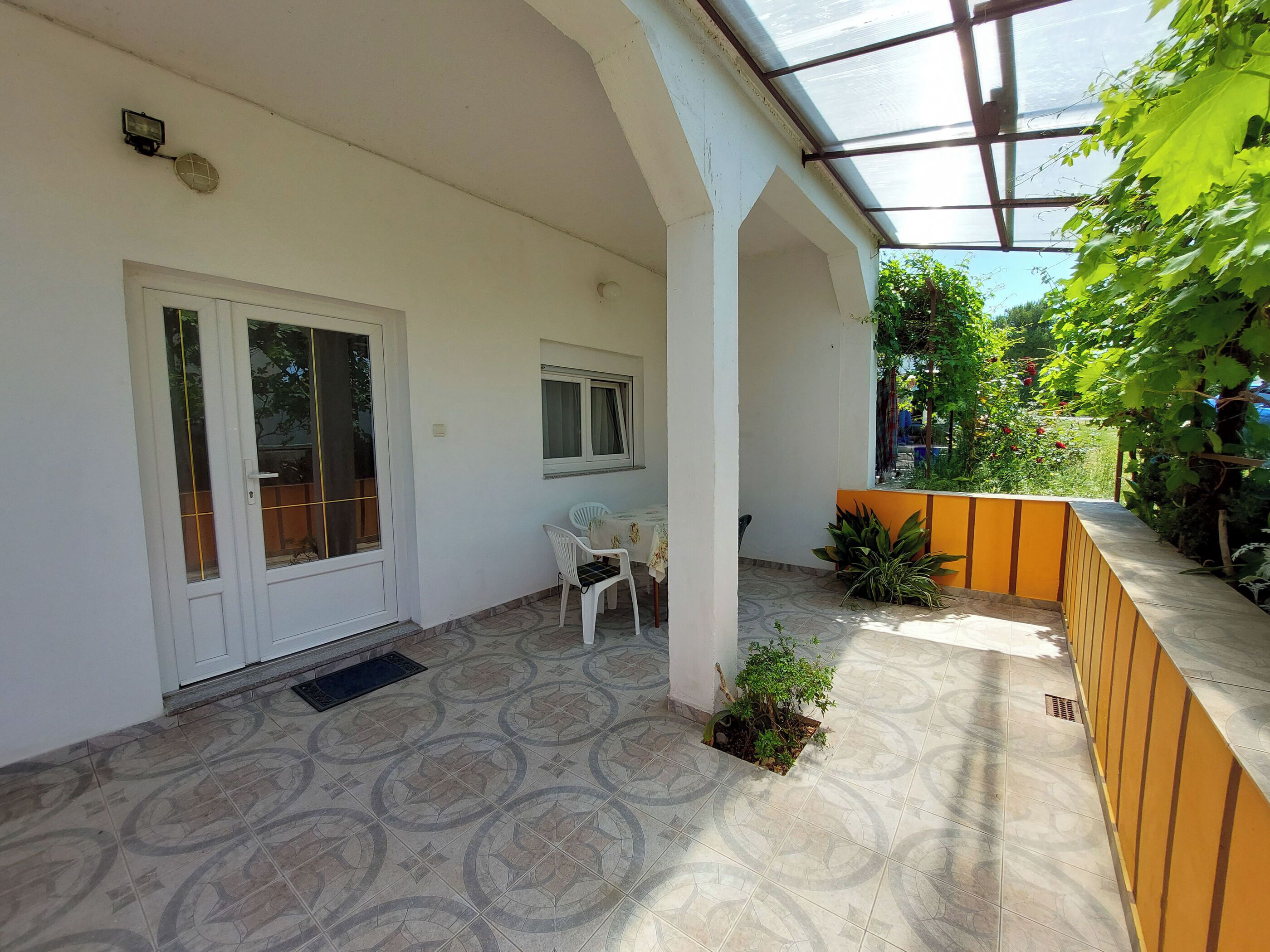 A covered patio area with a patterned tile floor, a white door, a small table with chairs, and potted plants.