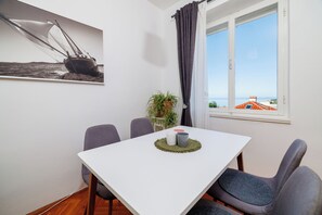 Une pièce avec une table blanche, quatre chaises grises, une plante en pot et une fenêtre donnant sur la mer et les bâtiments.