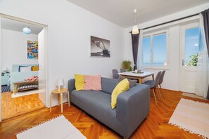 Un salon moderne avec un canapé gris, un plancher en bois, une table à manger avec des chaises, une petite table d’appoint et une vue sur la mer par une grande fenêtre.