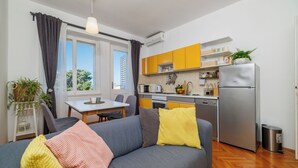 Eine moderne Wohnung mit einem grauen Sofa, gelb-rosa Kissen, einem Essbereich und einer Küche mit gelben Schränken sowie einem Edelstahl-Kühlschrank.