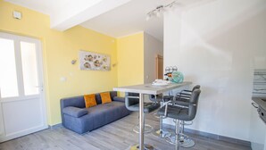 A modern kitchen with a grey countertop, a dining area with a grey sofa and two bar stools, and a yellow wall with a decorative piece. - Apartment Smile (Opatija)