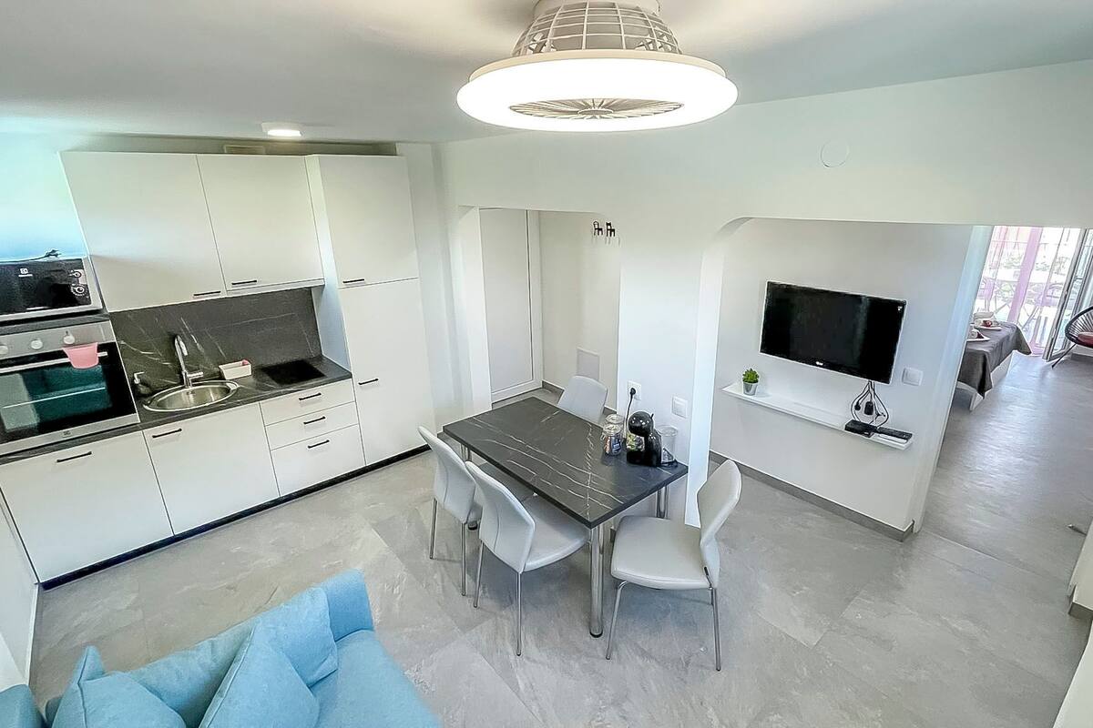 A modern kitchen with white cabinets, a built-in oven, and a dining area with a dark marble table and white chairs.
