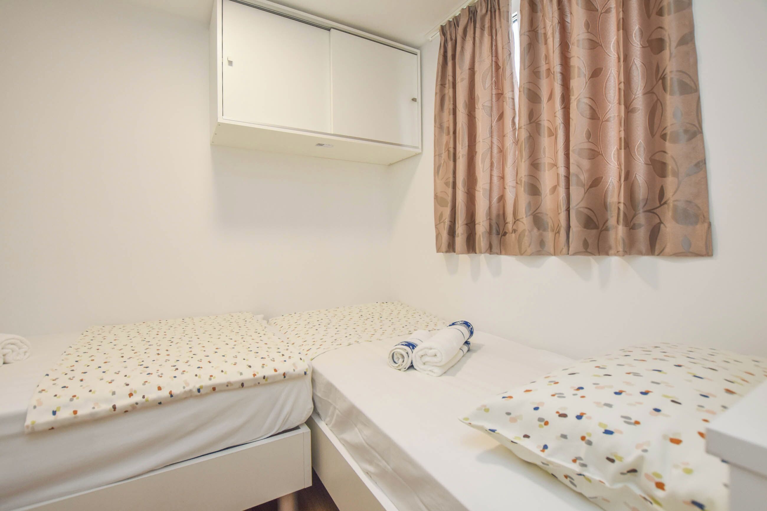 A small, clean room with two beds, a wardrobe, and a window with curtains.