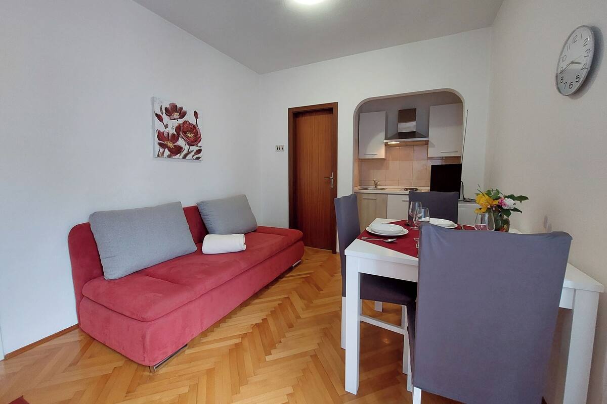 A compact living space with a red sofa, a dining table with chairs, a kitchen area, and a wall clock.