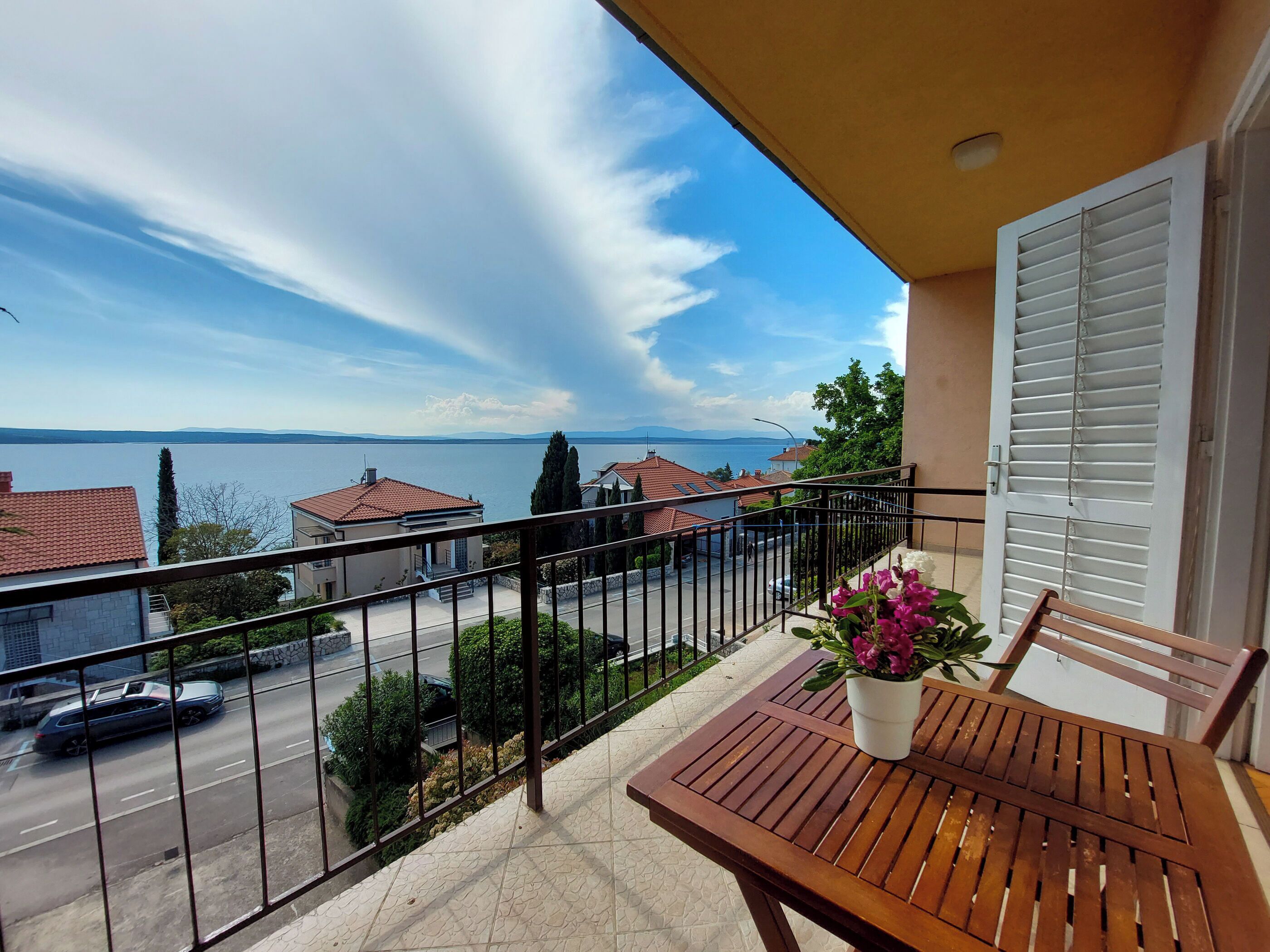 Ein Balkon mit Holztisch und Blumentopf, mit Blick auf eine Straße und ein Gewässer.