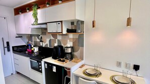 Egy modern konyha fehér szekrényekkel, beépített sütővel, mikrohullámú sütővel és kávéfőzővel. Van egy étkezősarok, egy két személyre megterített asztallal, egy székkel és egy mennyezetről lógó lámpával.