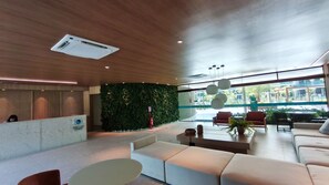 A modern lobby with a reception desk, seating area, and large windows overlooking a pool area. - Beach Class Verano - Porto de Galinhas (Ipojuca)