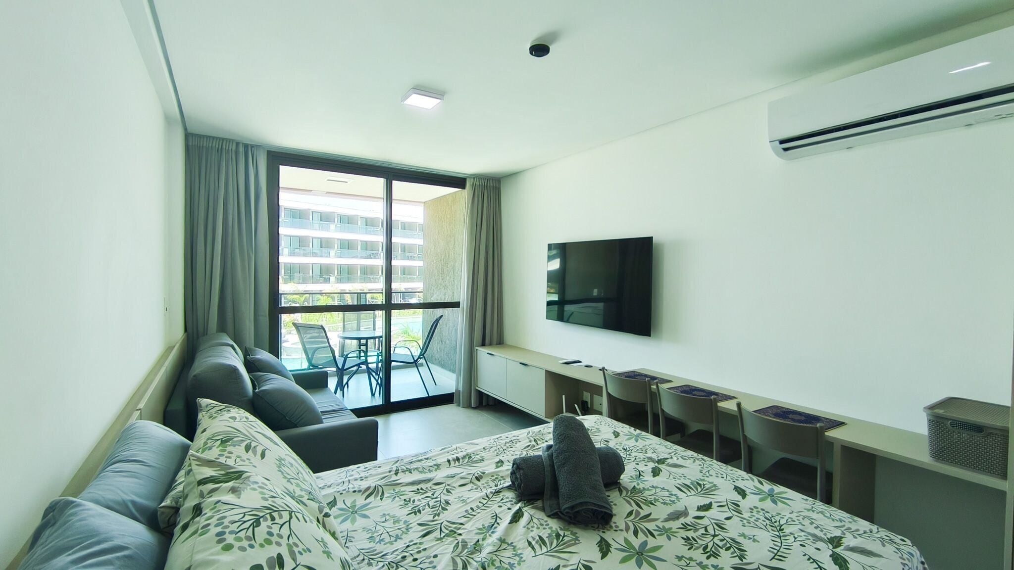 A modern hotel room with a bed, a TV, a balcony with chairs, and a desk.
