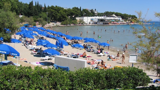 A beach with numerous blue umbrellas and sun loungers, a clear blue sky, and a view of the sea. - Apartment Astrid (Split)