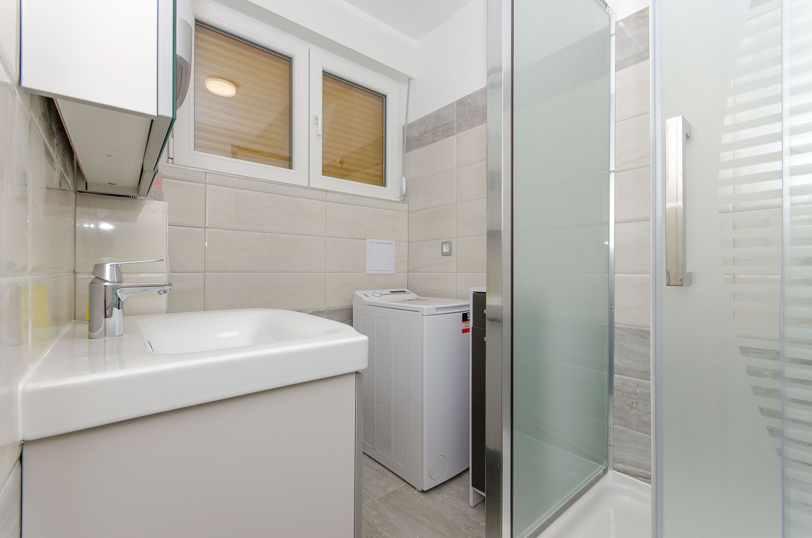 A modern bathroom with a sink, a washing machine, and a glass shower enclosure.