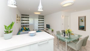 A modern kitchen with a white countertop, a dining area with a glass table, and a living area with a sofa and TV. - Apartment Astrid (Split)