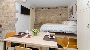 A cozy room with a stone wall, a bed with a white lace bedspread, a dining table with plates of food, and a TV mounted on the wall. - Apartment Pavlimir (Solta)