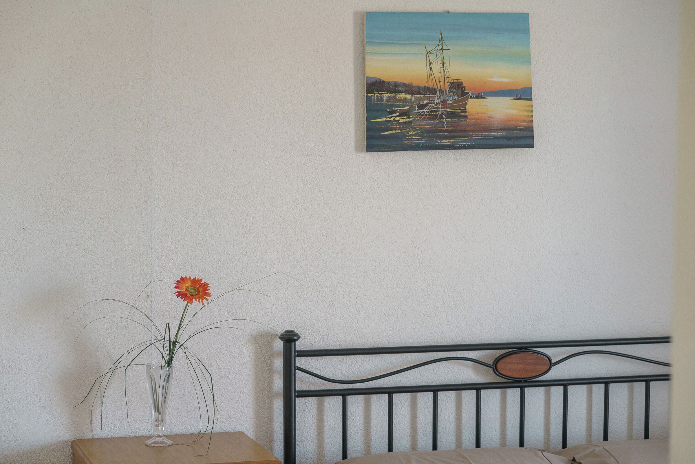 Une chambre à coucher avec un lit, une table de chevet ornée d’un vase de fleurs et un tableau représentant un coucher de soleil sur l’eau, accroché au mur.