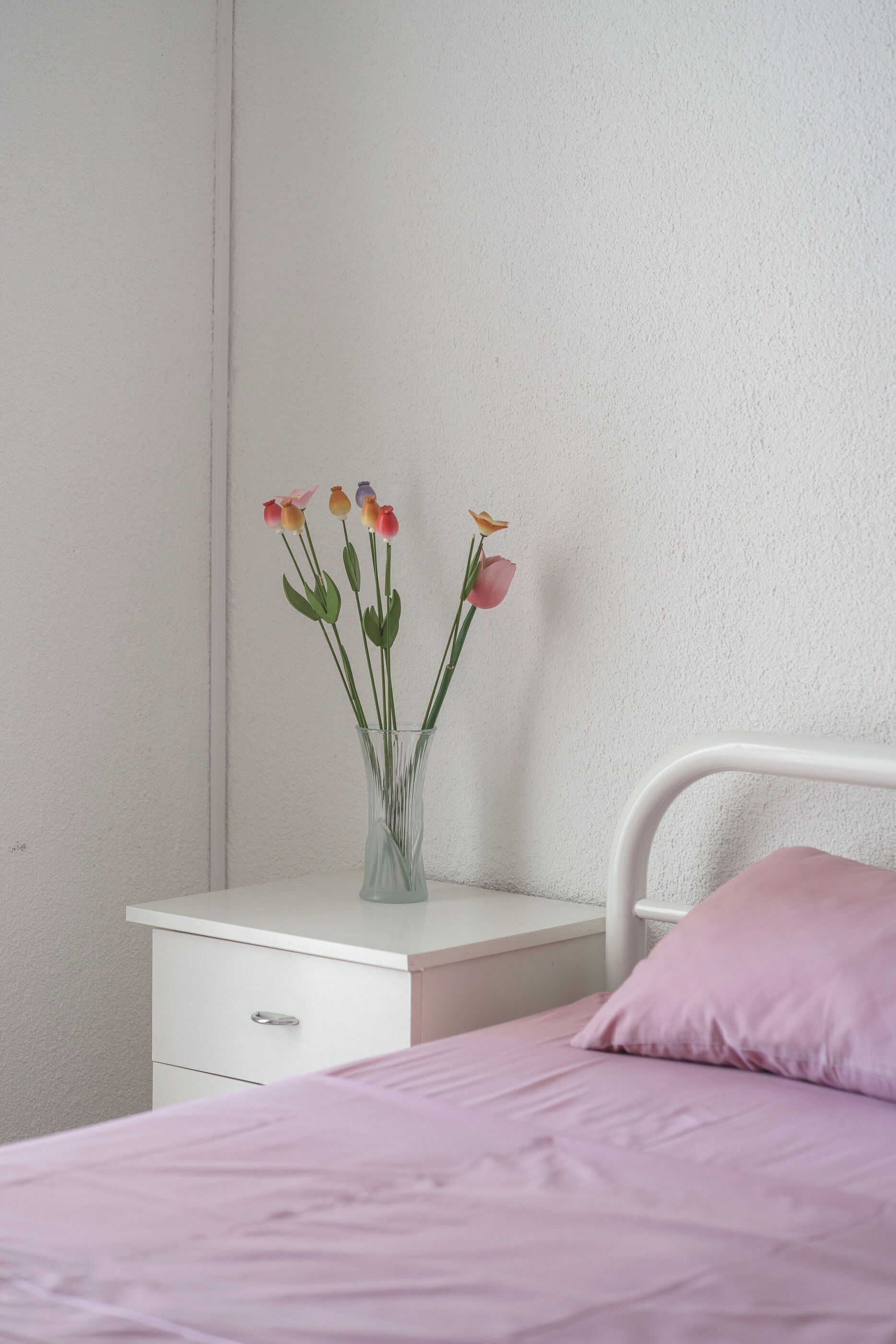 Een slaapkamer met een bed, een nachtkastje, een vaas met bloemen en een witte muur.