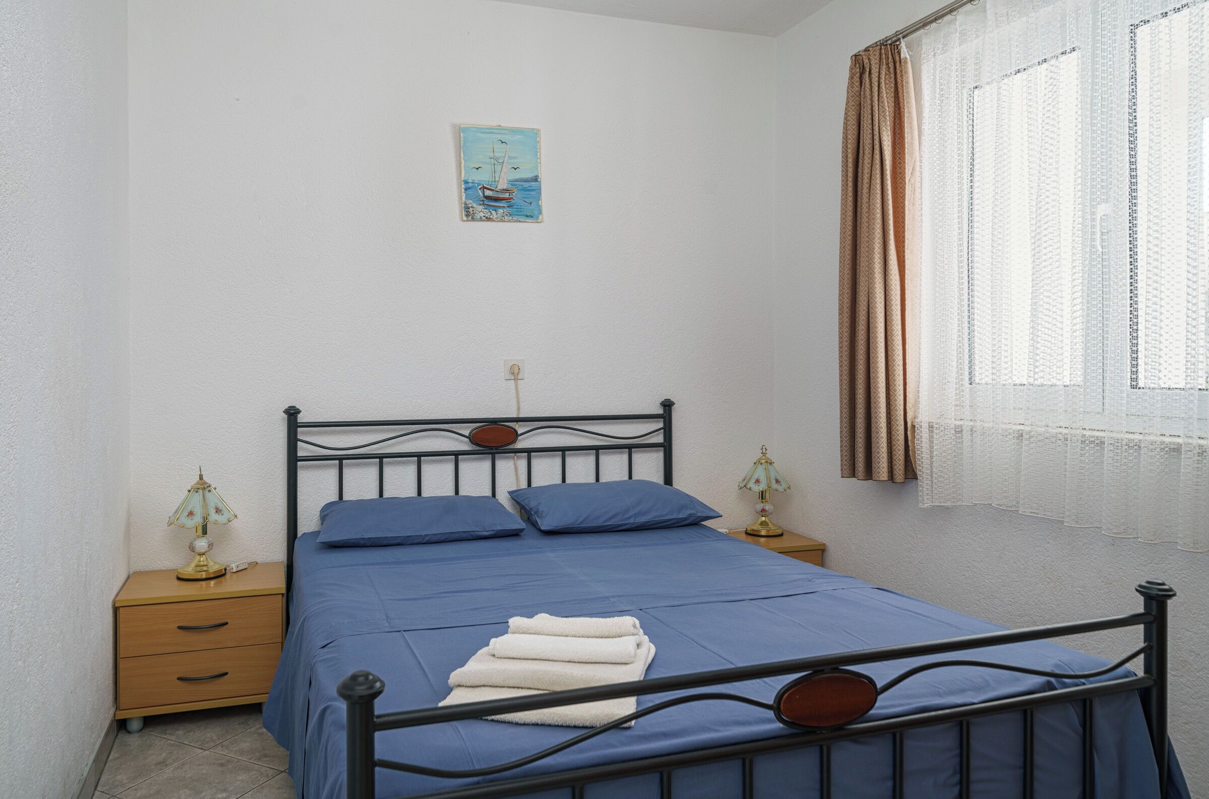 A single bed with a metal frame, two bedside tables with lamps, a blue bedspread, and a painting on the wall.